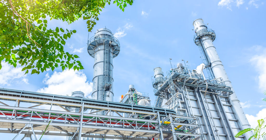 Sunlight shines through green leaves on an industrial gas plant with tall towers.