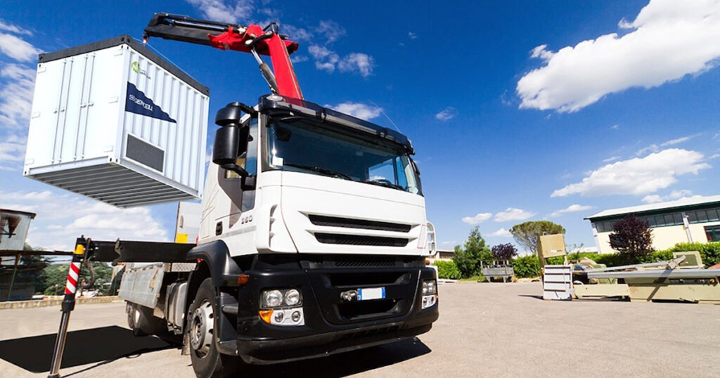 Truck with crane lifting a Gencell shipping container in a sunny outdoor area.