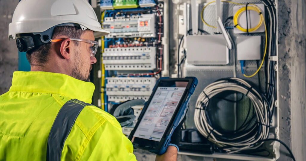 Electrician with hard hat using a tablet to inspect an industrial control panel.