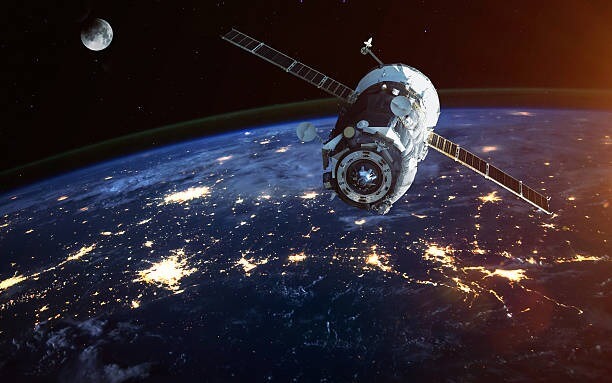 Image of satellite above the earth, with illuminated areas on the earth showing activity, with the moon in the background