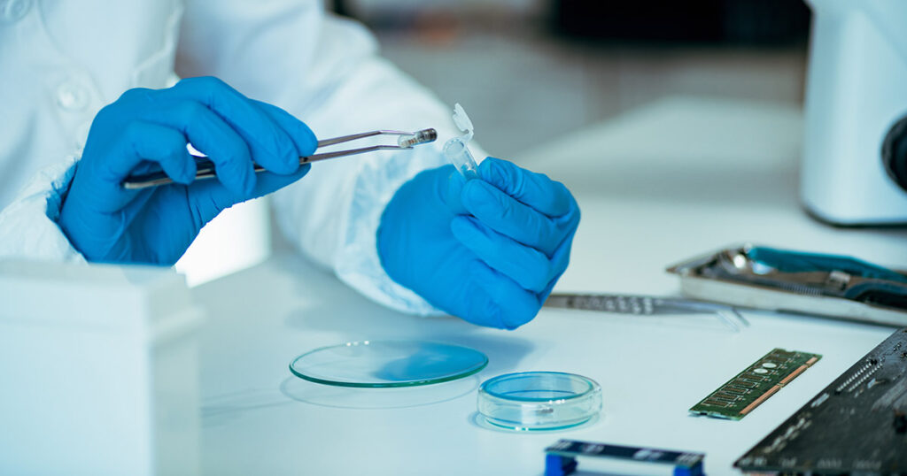 Lab technician in blue gloves handling a sample with tweezers in a bright laboratory setting.