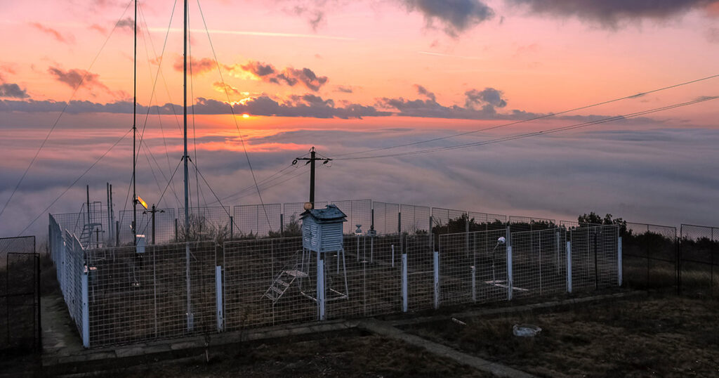 A weather station at sunset with vibrant clouds and antennas silhouetted.