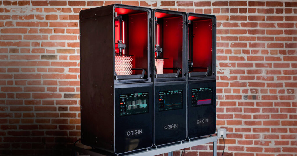 Three Origin 3D printers creating objects with red illumination in a room with a brick wall.